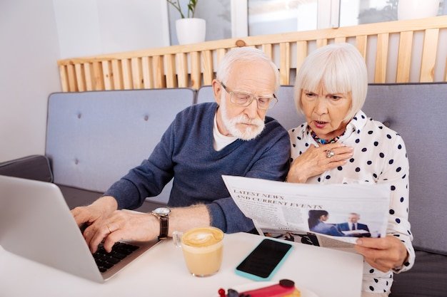 Concerned senior couple at a cafe reviewing a newspaper and typing on a laptop, working to avoid double taxation on retirement income