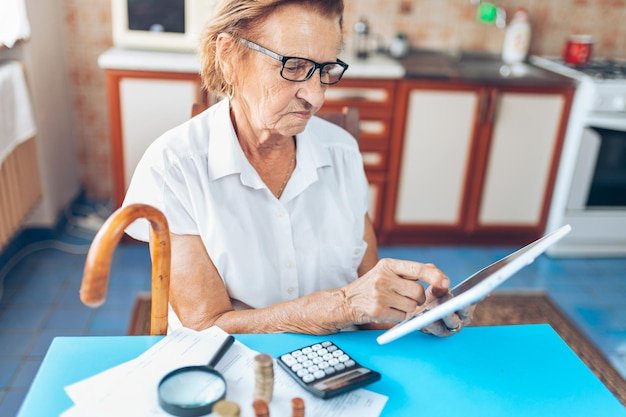 Older woman using a tablet with a calculator and documents at a kitchen table calculating her Social Security break-even age