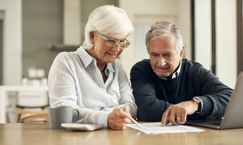 Elderly couple reviewing and signing documents at a kitchen table with a laptop, checking beneficiary designations correctly