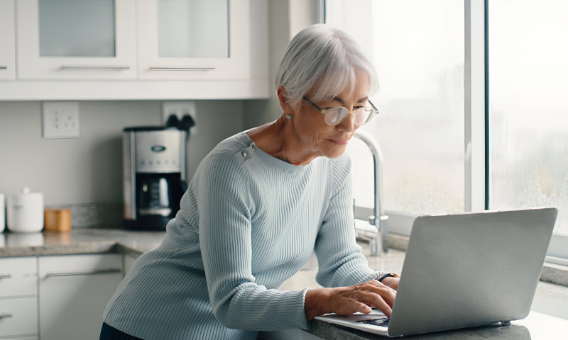 Senior woman with glasses on a laptop in a bright kitchen managing combined Social Security and pension income