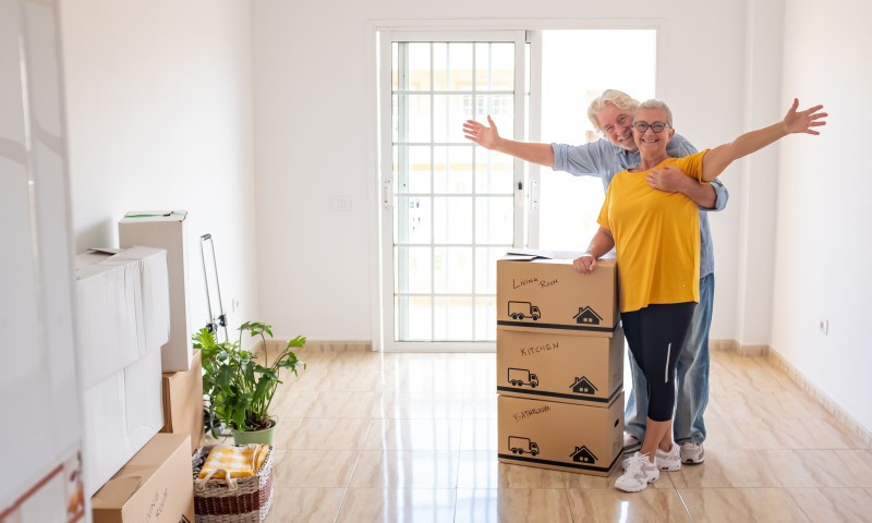 Senior couple surrounded by labeled moving boxes in a bright apartment, thoughtfully downsizing for retirement