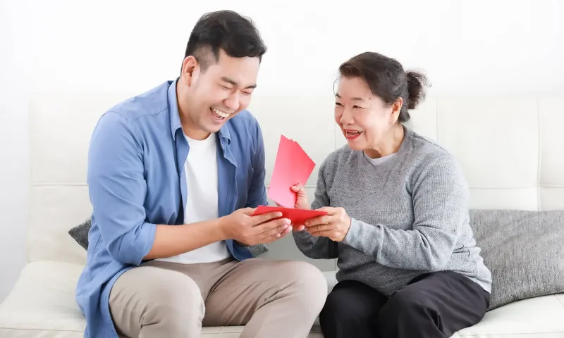 Smiling mother giving her adult son a red gift card on a sofa, illustrating how retirees legally gift money to family members