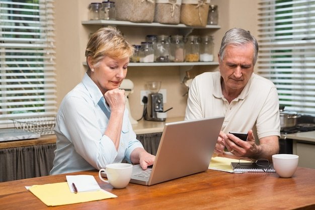 Middle-aged couple at a kitchen table with a laptop and phone, plotting mini-retirements to experience freedom in their 30s and 40s