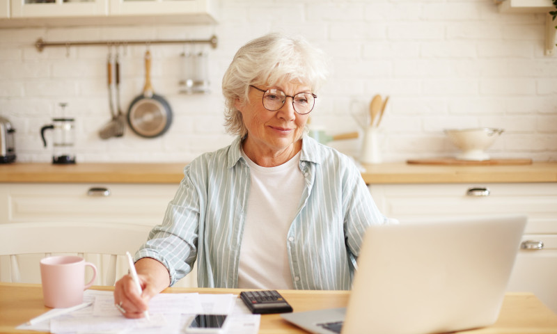 Senior woman writing retirement income notes beside a laptop and calculator at her kitchen table