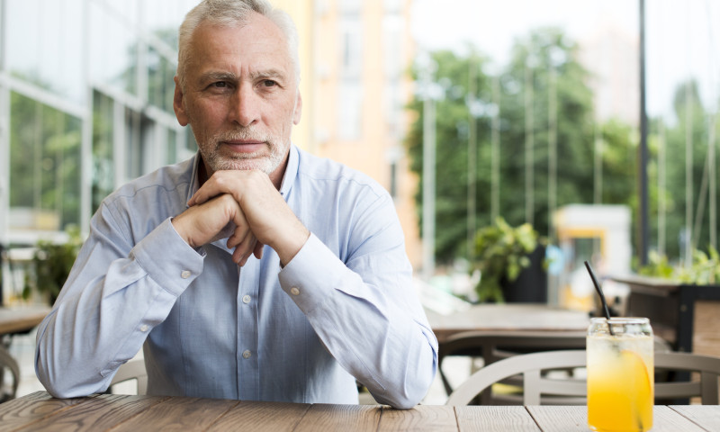 Thoughtful senior man sitting at an outdoor cafe table with a juice, contemplating whether he is ready to retire early