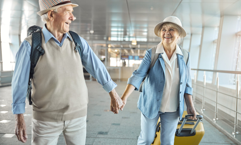 Senior couple holding hands walking through an airport terminal with luggage, exploring how moving abroad affects Social Security