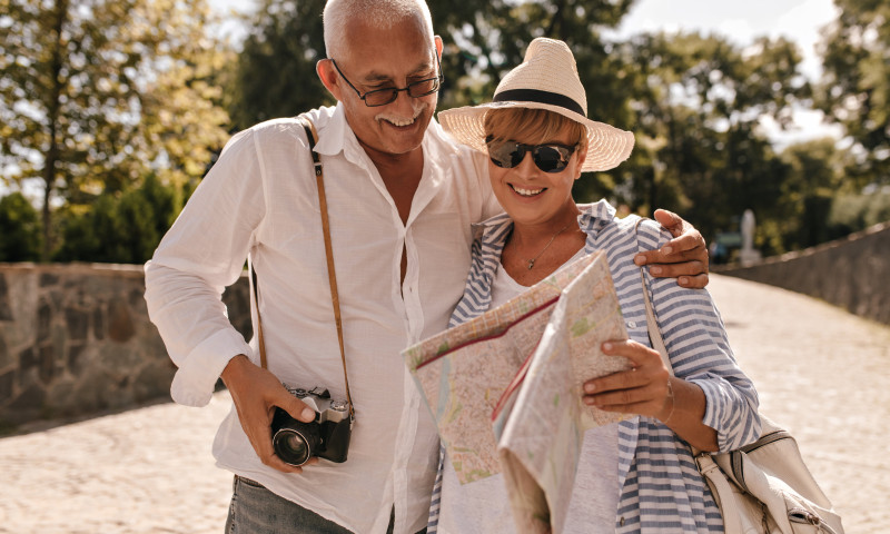Senior couple smiling while reading a travel map outdoors with a camera, budgeting for travel expenses in retirement