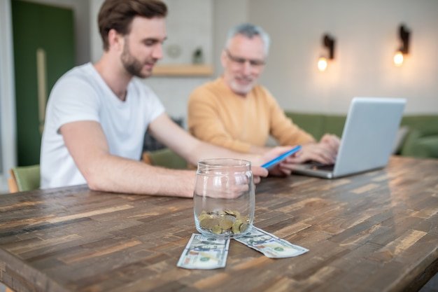 Two men at a table with a jar and dollar bills, discussing how 401k employer matching is free retirement money