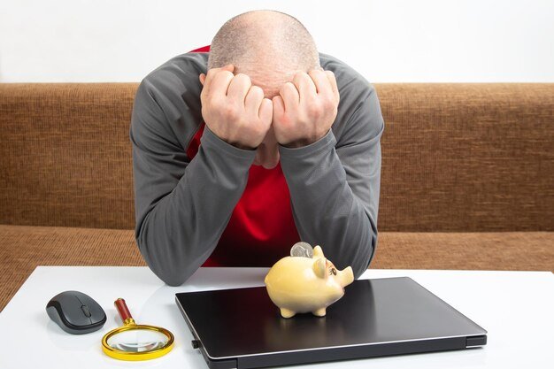 Man with head in hands in front of a piggy bank and laptop, illustrating the financial consequences of cashing out a 401(k)