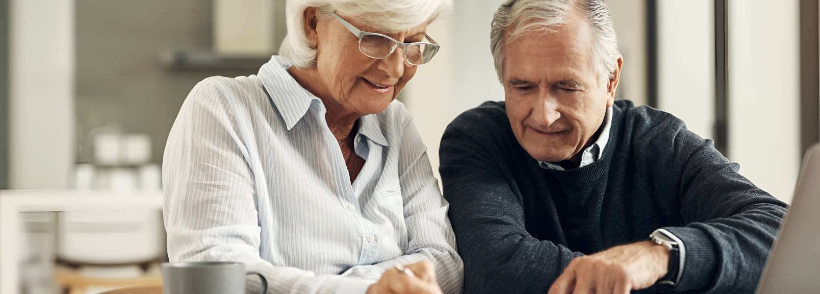 Elderly couple reviewing and signing documents at a kitchen table with a laptop, checking beneficiary designations correctly