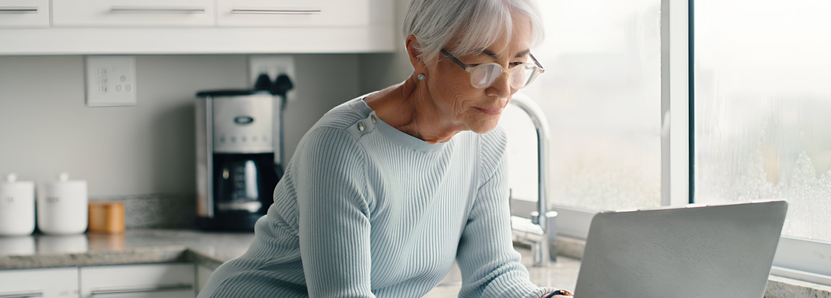 Senior woman with glasses on a laptop in a bright kitchen managing combined Social Security and pension income