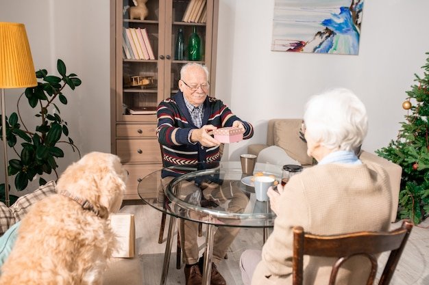 Elderly man sharing a gift with another senior while a dog sits nearby in a cozy co-living home