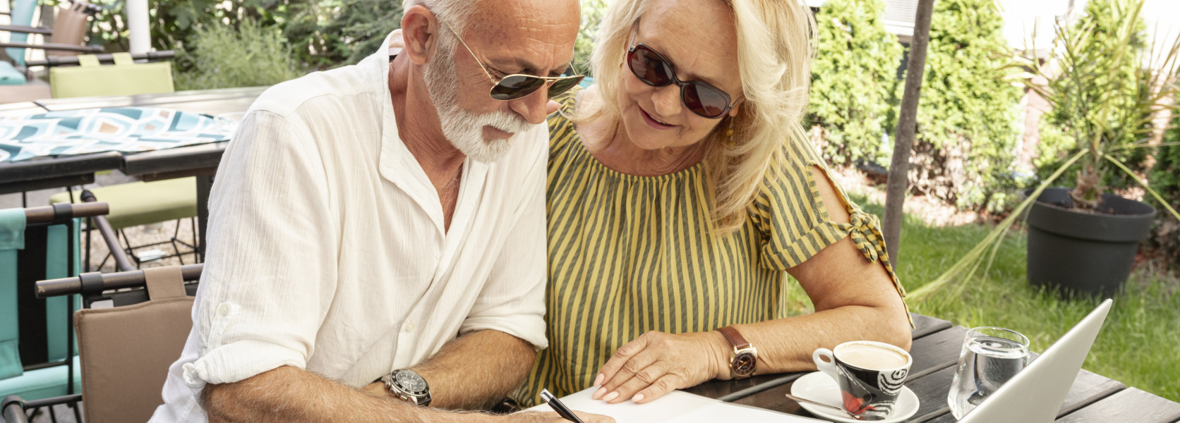 Senior couple reviewing retirement paperwork on a laptop at an outdoor cafe, planning their final 5 years before retiring
