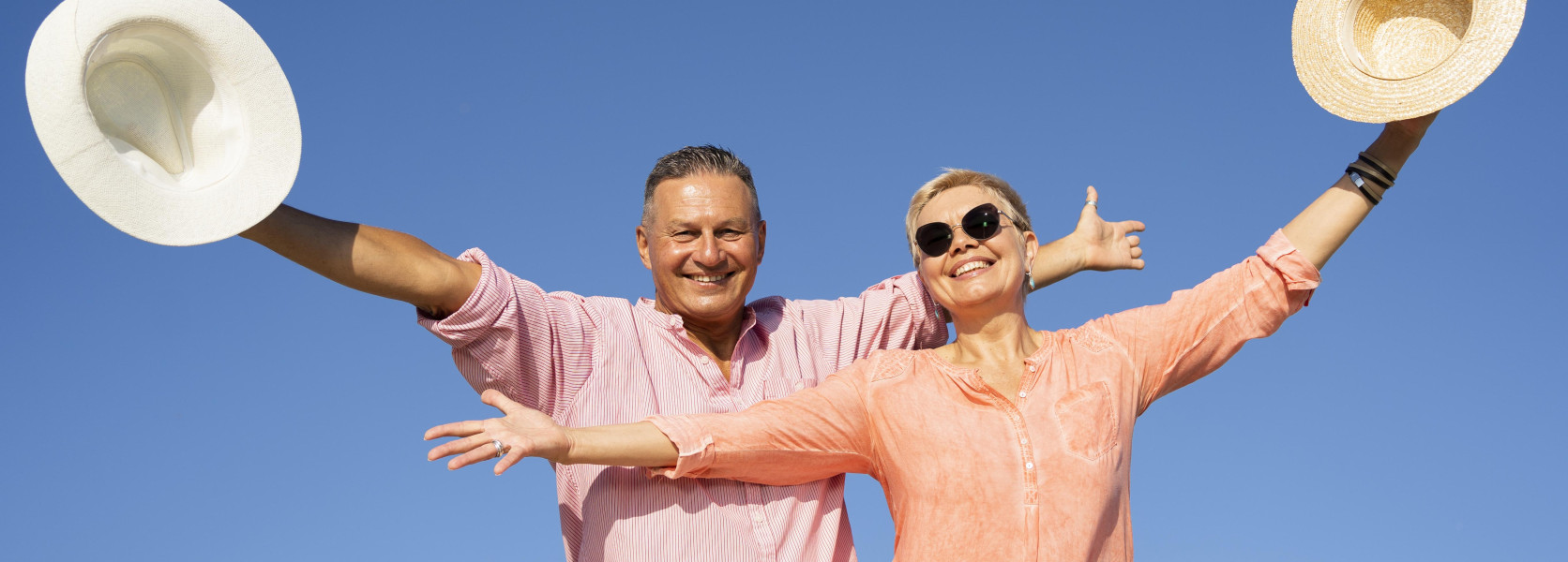 Older couple raising straw hats outdoors, celebrating the freedom of a comfortable retirement