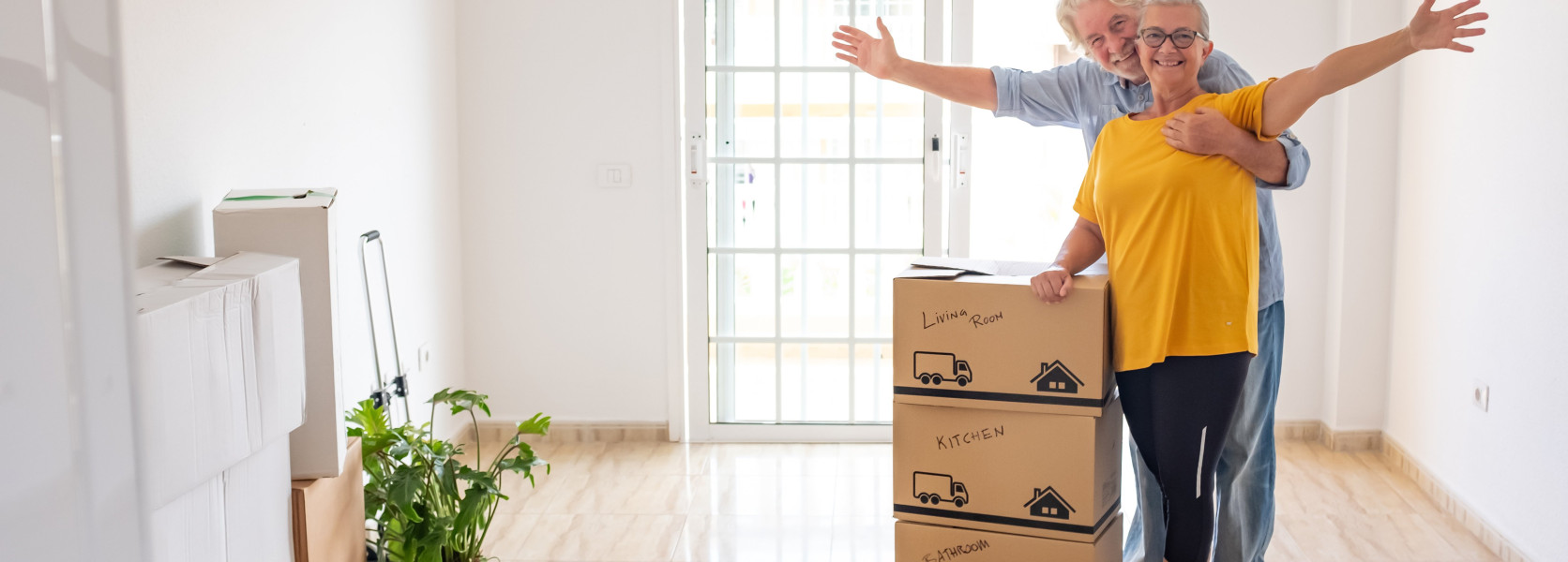 Senior couple surrounded by labeled moving boxes in a bright apartment, thoughtfully downsizing for retirement