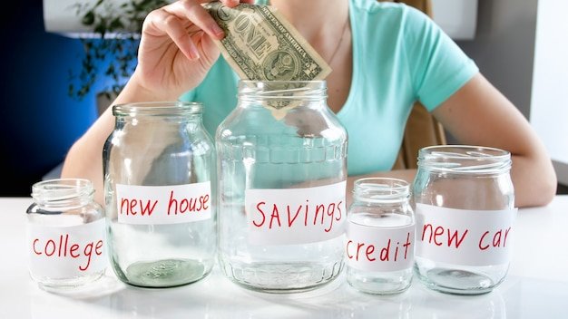 Person inserting a dollar bill into a Savings jar beside jars for college and other goals, maximizing 401(k) contributions
