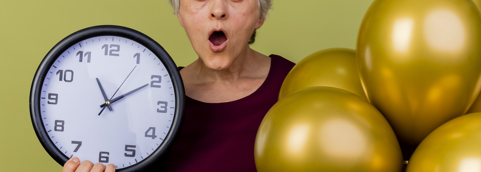 Older woman holding a wall clock with gold balloons, representing the decision to delay Social Security claiming until age 70