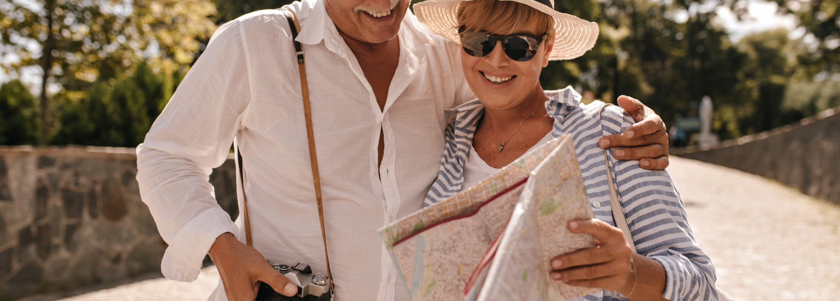 Senior couple smiling while reading a travel map outdoors with a camera, budgeting for travel expenses in retirement
